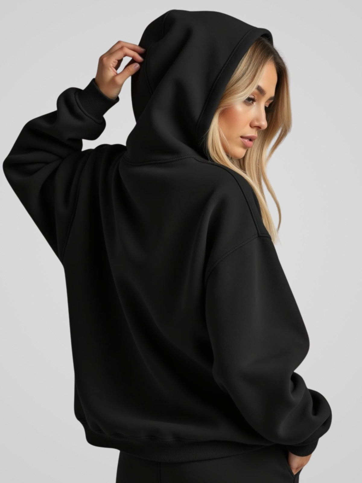Woman wearing a black oversized hoodie with the hood up, standing backwards and facing forward to show the fit and style.